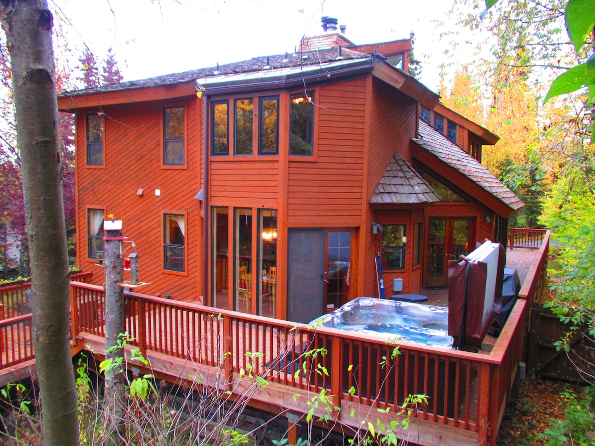 The charming exterior of a three-story wood-framed house is surrounded by greenery, showcasing large windows that provide ample natural light. A spacious deck features a hot tub and is framed by wooden railings, inviting relaxation amidst the natural surroundings.
