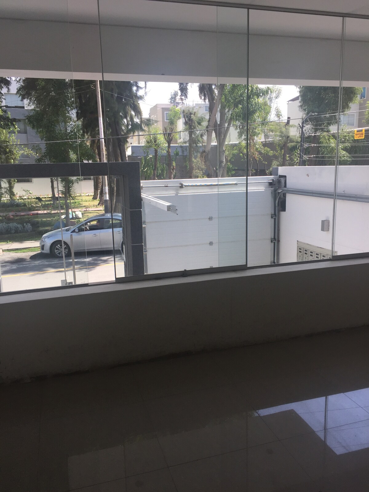 A large window provides a view of the street and surrounding greenery. A parked gray vehicle can be seen, alongside trees lining the area. The interior space features glossy tile flooring that reflects light, enhancing the overall brightness of the room.