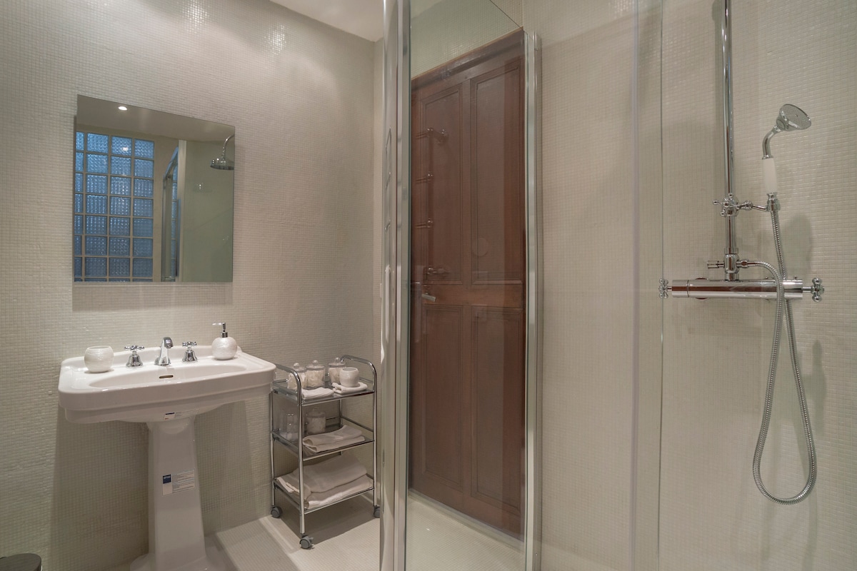 The bathroom features a spacious walk-in shower area, equipped with modern fixtures. A stylish sink is positioned beside a mobile cart with towels and toiletries. Natural light filters through a frosted window, illuminating the elegant, minimalist design.