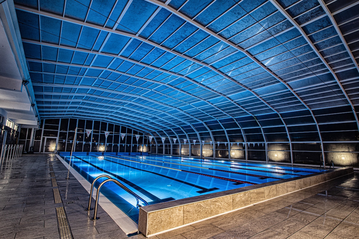 An indoor swimming pool is shown under a high, transparent roof, allowing natural light to filter through. The clear waters of the pool are illuminated with soft blue lighting, and diving lanes are marked along the length of the pool. The surrounding area features smooth tile flooring.