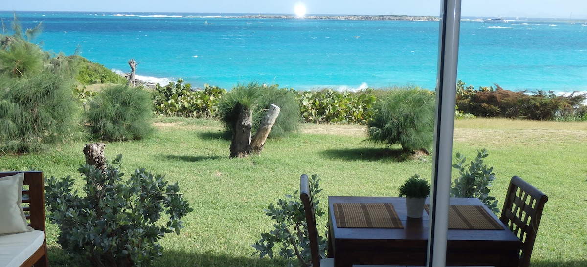 A dining area is set outside, featuring a wooden table with seating for four. Lush greenery surrounds the space, offering a view of the turquoise ocean beyond. The scene highlights the tranquil beach atmosphere, with gentle waves visible in the distance.