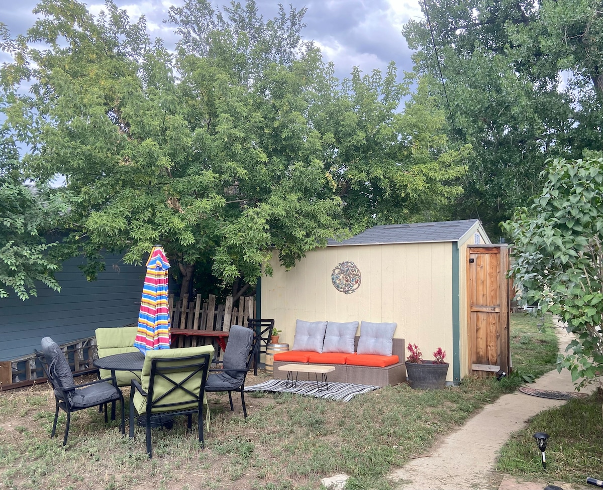 A private backyard space is shown, featuring a patio area with a seating arrangement. A table with chairs sits beside a lounge area covered by a colorful umbrella. A small storage shed is visible against the backdrop of greenery, contributing to a relaxed outdoor atmosphere.