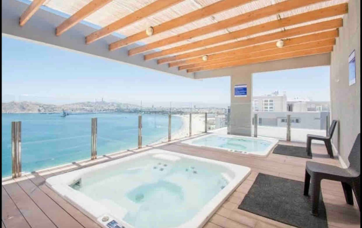 Two outdoor jacuzzis are positioned on a wooden deck, offering views of the sea. A pergola with exposed beams provides shade above. A single chair and a small mat are placed nearby, creating a relaxed space for enjoyment while overlooking the water.