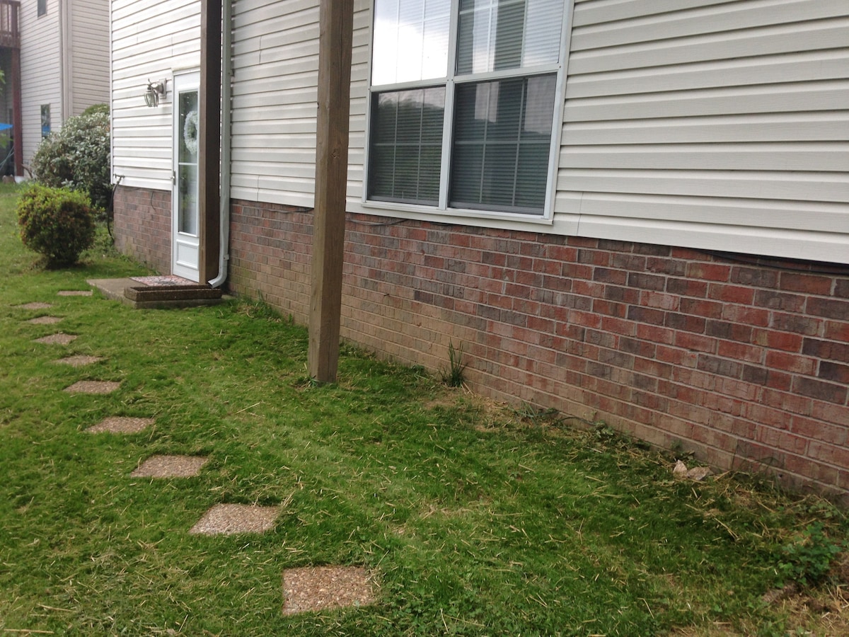 A well-maintained outdoor area features a grassy lawn with stepping stones leading up to a private entrance. The exterior is characterized by light-colored siding and a brick foundation, with ample natural light coming through the nearby windows.