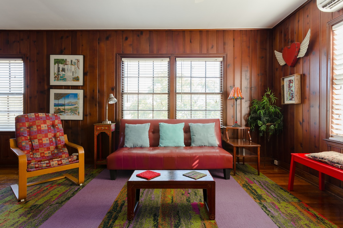 The living area features wooden paneling and multiple windows allowing natural light to brighten the space. A red sofa with cushions faces a low coffee table, complemented by colorful chairs and decor elements, including framed art and a leafy plant.