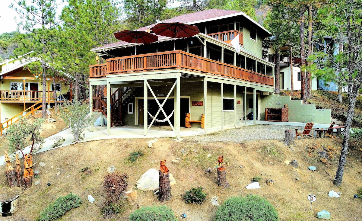 The exterior of the lake house features a multi-level structure surrounded by trees. A spacious deck with umbrellas provides outdoor seating. The property includes a gravel area with a fire pit and decorative wooden carvings on the hillside, set against a natural landscape.