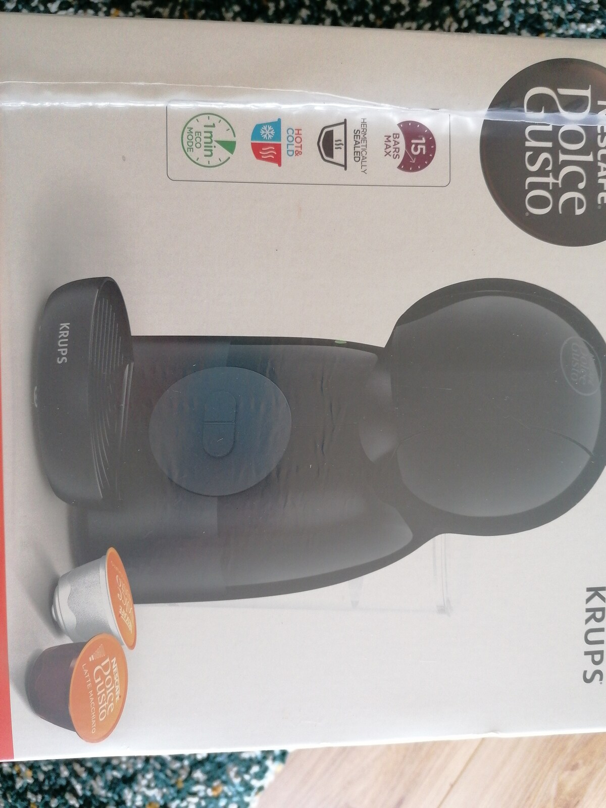 A sleek coffee machine is displayed on its packaging, featuring a modern black design with a central button. Two coffee capsules are positioned next to the machine, showcasing different colors and branding. The overall presentation emphasizes a contemporary kitchen appliance.