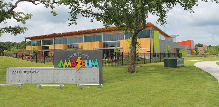 The exterior of the Scott Family Amazeum is shown, featuring a modern design with colorful panels and large glass windows. The building is surrounded by a manicured lawn and walking paths, emphasizing the spacious outdoor area. A clear sign marks the entrance to the museum.