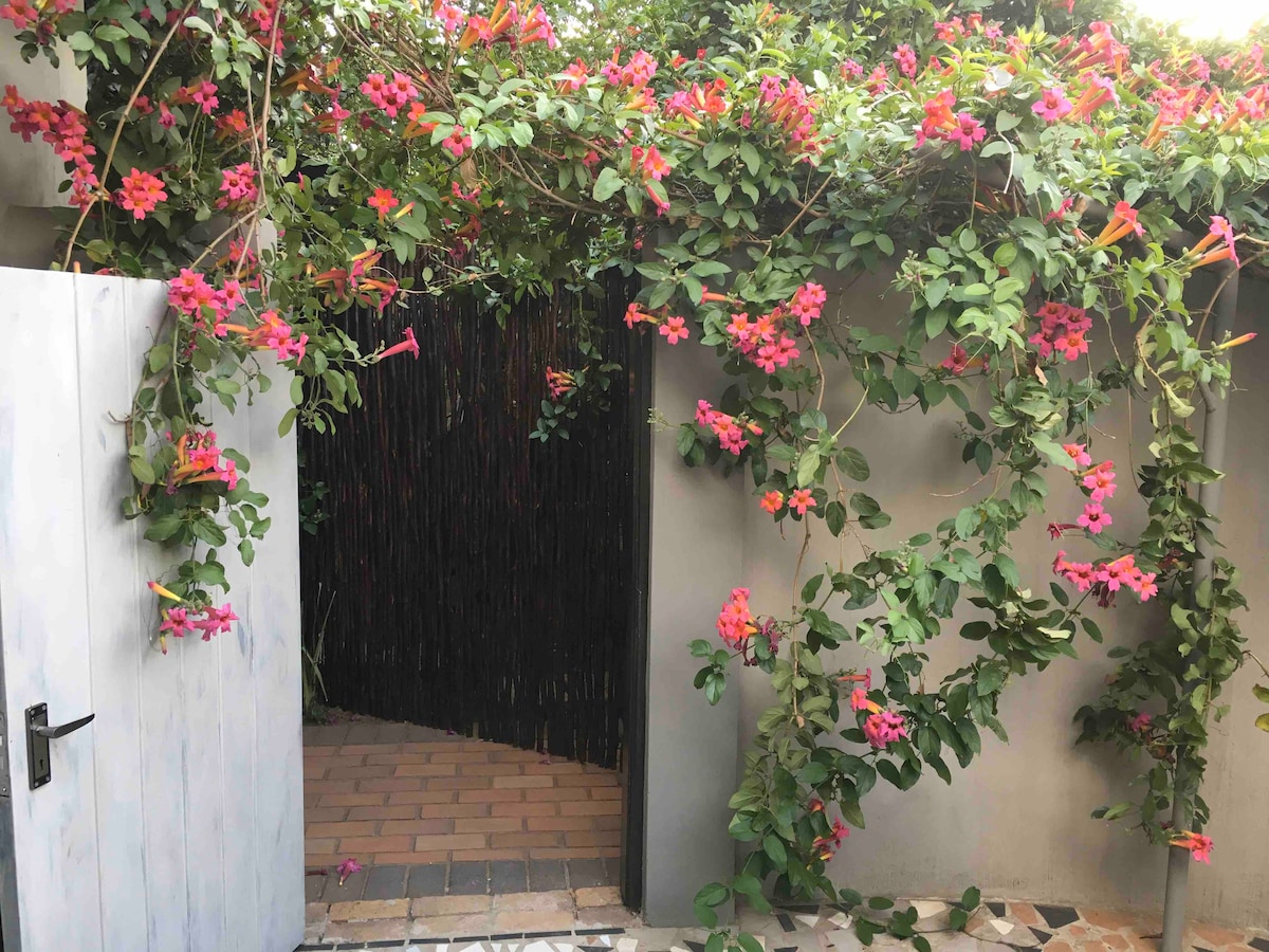 A vibrant entrance is framed by lush flowering vines, featuring pink blossoms cascading from above. A white wooden gate is positioned on one side, revealing a brick pathway leading into a serene outdoor area surrounded by natural textures.