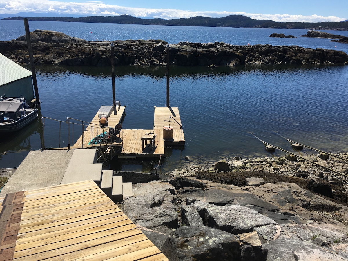 A wooden dock extends into calm waters, bordered by rocky shoreline. Various boats are visible at the dock, and a small structure is positioned nearby. The landscape features distant hills under a clear sky, reflecting a serene waterfront atmosphere.