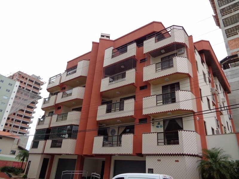 The building features a modern architecture with a red and cream facade, structured in several levels. Multiple balconies are visible, each with black metal railings. Ground-level parking spaces are provided, along with well-maintained greenery flanking the entrance.