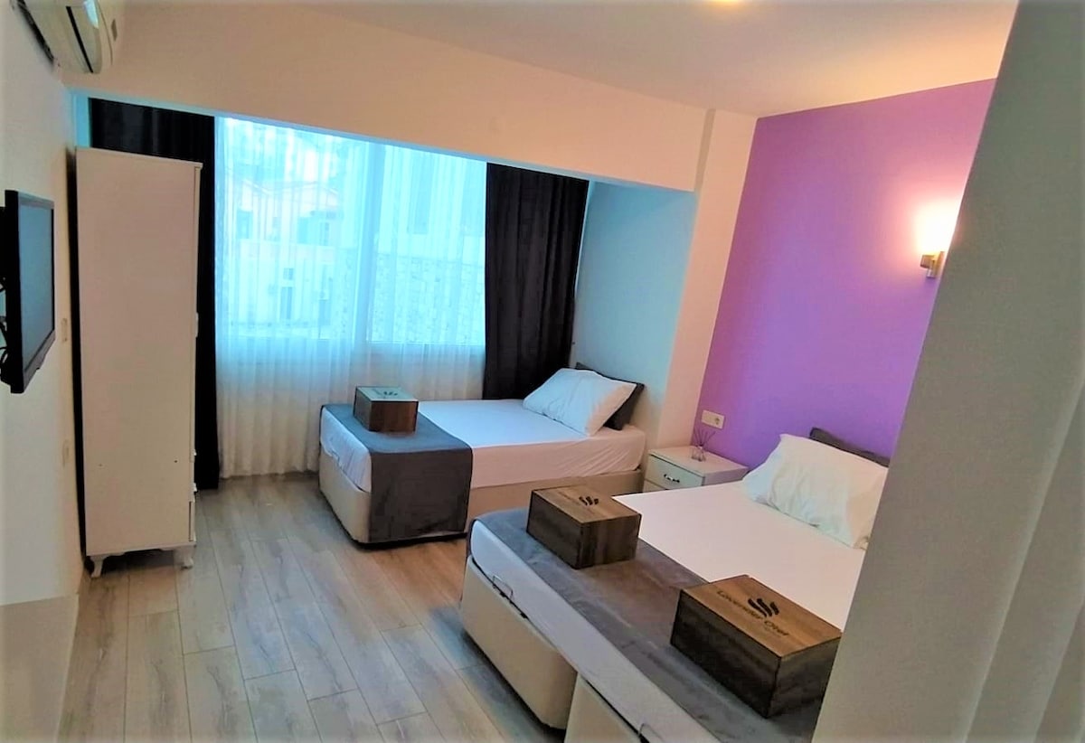 A family room features a double bed and a single bed, both dressed in white linens. A large window allows natural light to fill the space, with sheer curtains softly diffusing the light. Nightstands are positioned beside each bed, providing additional functionality.