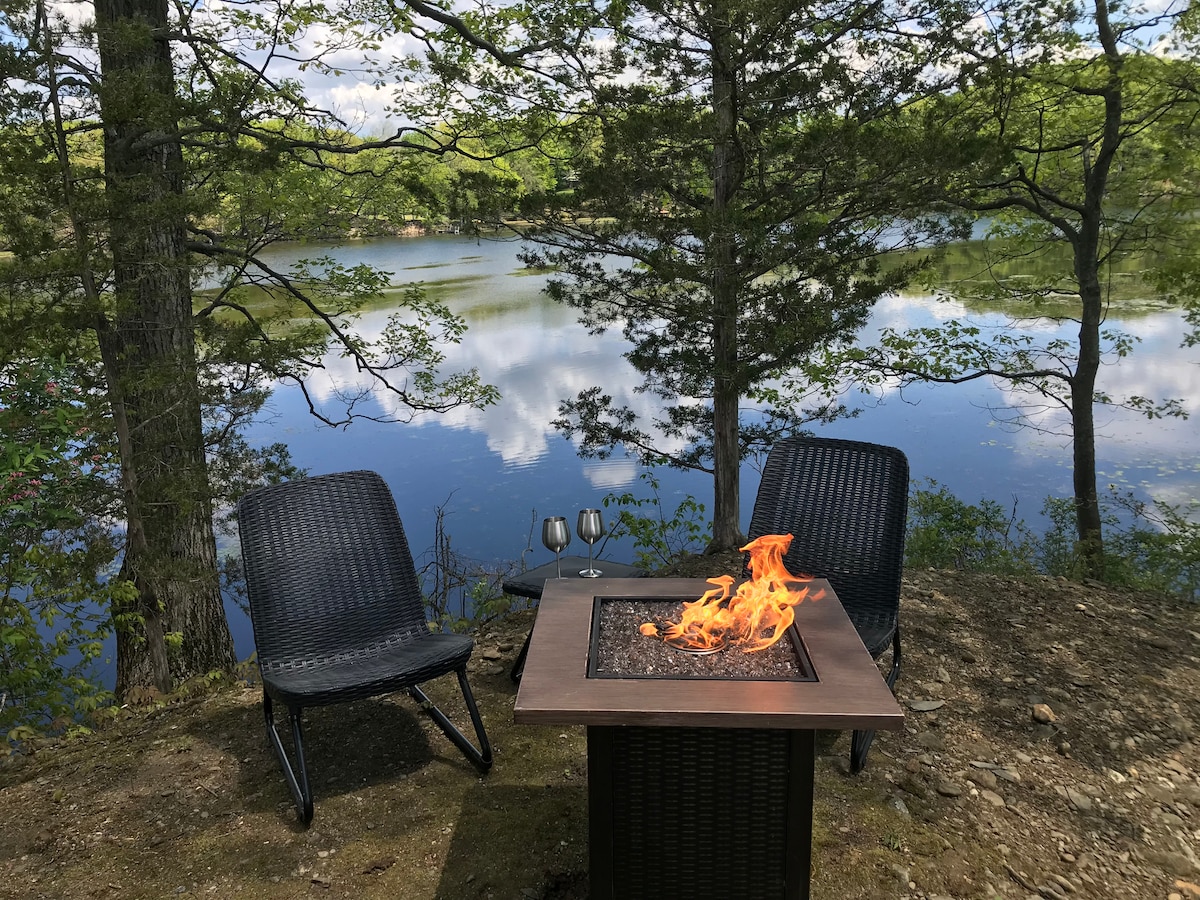 Two outdoor chairs are positioned near a fire pit, with flames flickering above a stone surface. A serene lake is visible in the background, surrounded by trees, reflecting the sky. Two glasses are placed on a small table nearby.