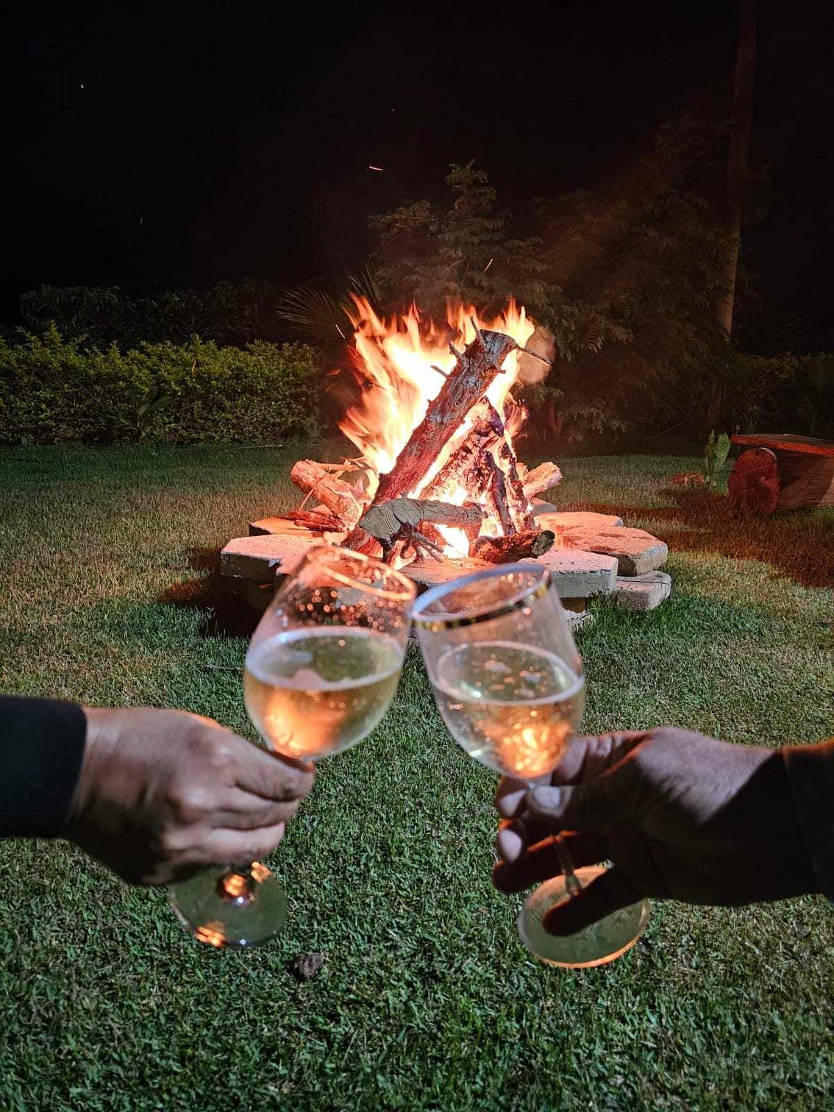 Two glasses of bubbly are held in front of a glowing bonfire surrounded by a stone circle. In the background, a grassy area is illuminated by the firelight, while foliage adds a natural touch to the serene nighttime setting.