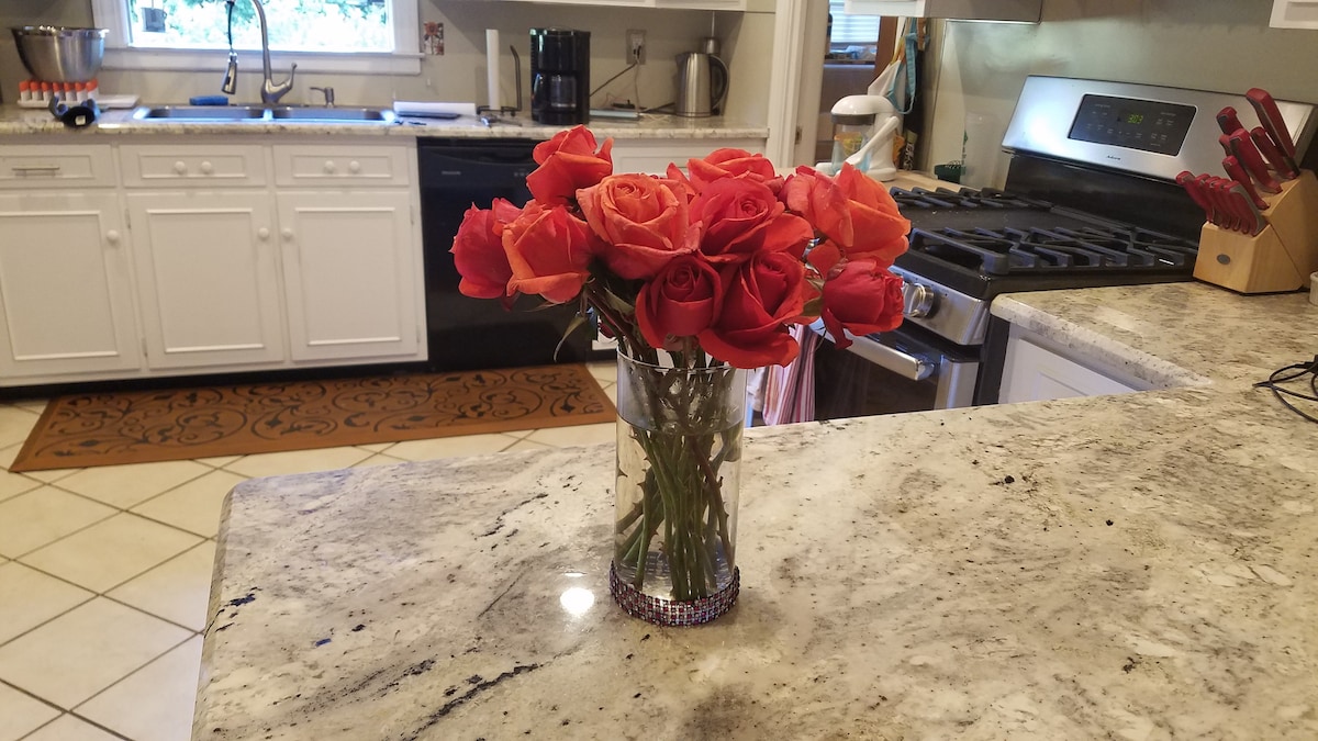 A vase filled with vibrant red and orange roses is centered on a granite countertop. The modern kitchen features white cabinets, a stainless steel stove, and a variety of appliances in the background. Natural light enters through a nearby window, enhancing the room's brightness.