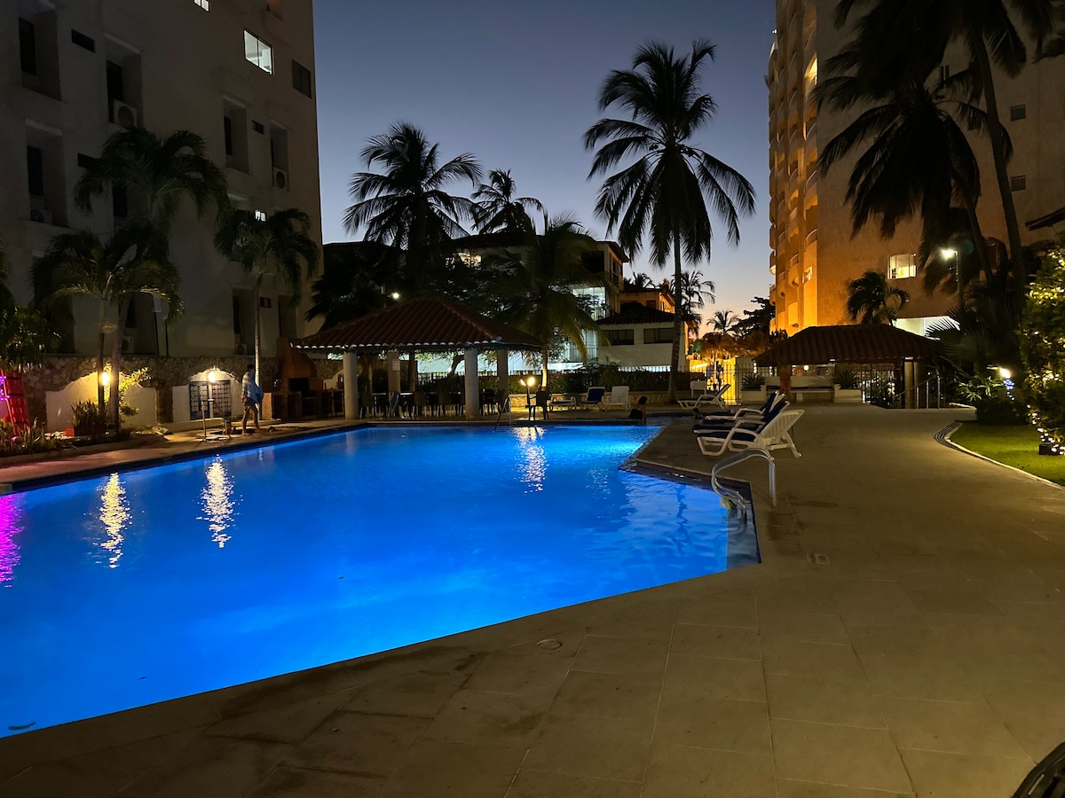 An illuminated pool reflects soft blue light under a twilight sky, surrounded by palm trees. Lounge chairs are positioned nearby, offering seating options. A thatched-roof gazebo is visible in the background, contributing to the laid-back atmosphere of the outdoor space.