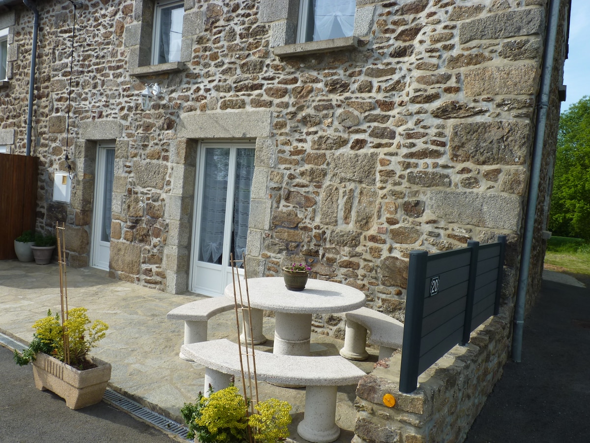 The exterior of the stone house is displayed, featuring a cozy seating area with a circular table and four stone stools. Potted plants add a touch of greenery, while large windows allow for natural light. A slate grey fence partially encloses the space for privacy.