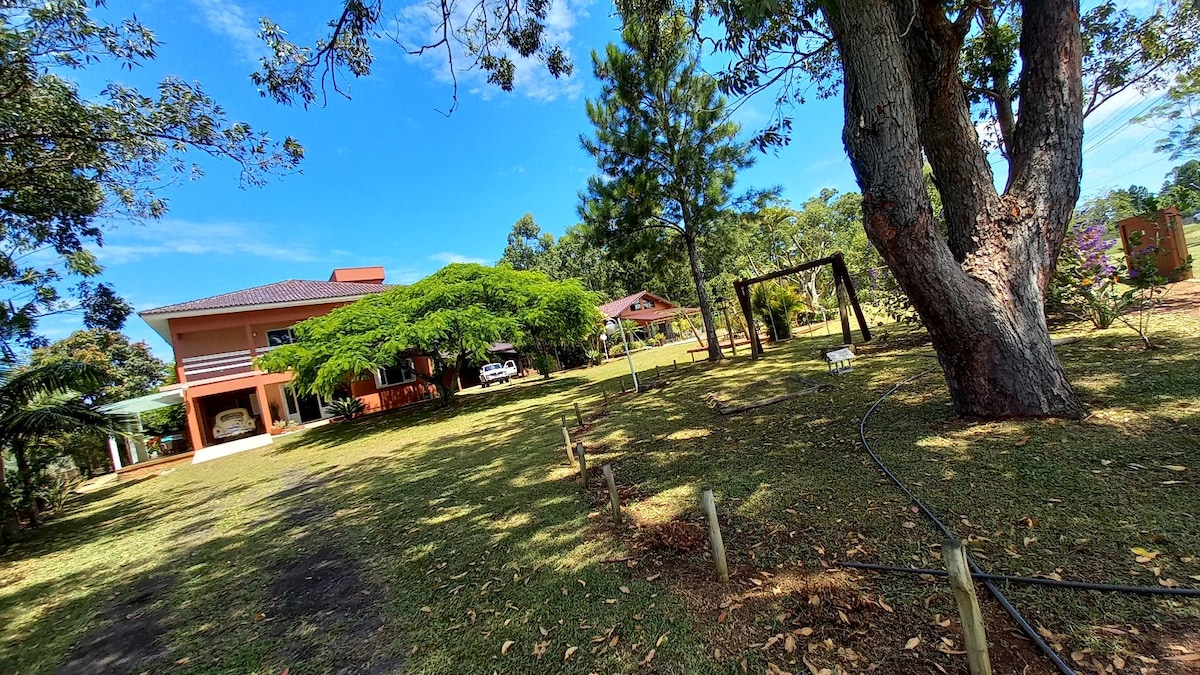 A spacious outdoor area is surrounded by lush greenery, featuring a large tree and a swing set. The two-story house sits in the background, with a gazebo visible nearby. Open grounds invite relaxation, while a clear blue sky complements the tranquil setting.
