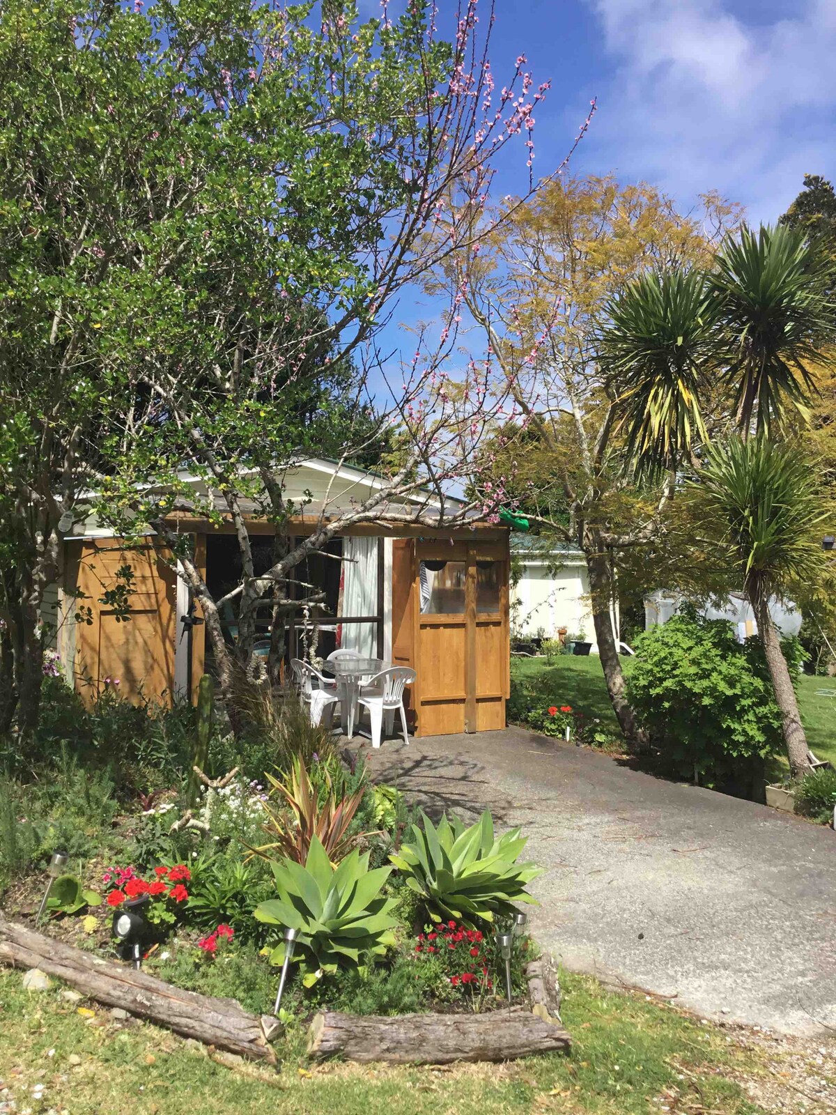 The cabin entrance is framed by vibrant greenery and blooming flowers. A gravel path leads to the wooden doors, flanked by a couple of white chairs. Trees and plants provide shade, enhancing the natural setting that surrounds the cabin.