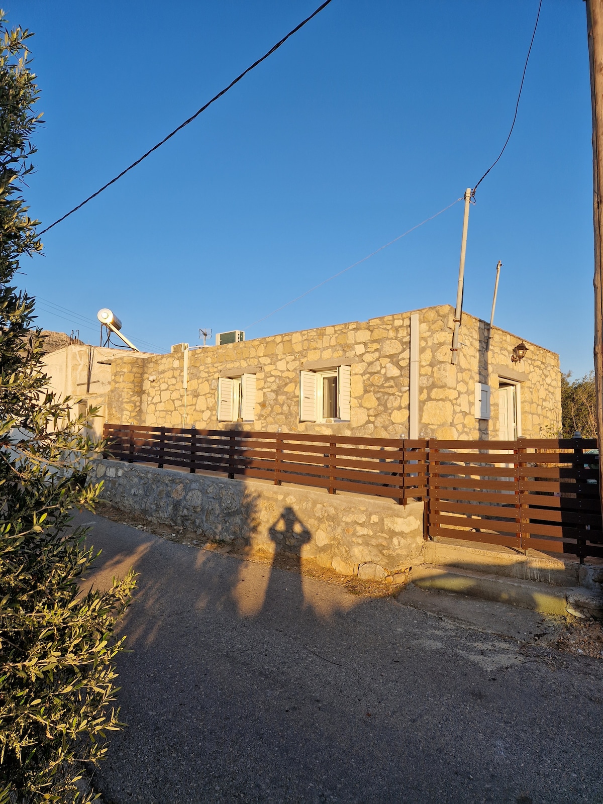 A rustic stone house is set against a clear blue sky, showcasing its traditional architecture. A wooden fence lines the property, and shadows cast by trees and utility poles add depth to the scene. The surrounding area appears peaceful and inviting.