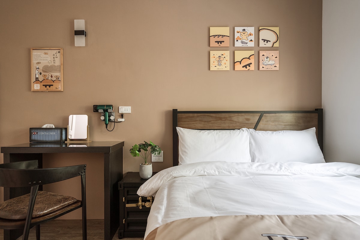 A cozy sleeping area features a spacious bed with a white comforter and a wooden headboard. A small desk with a chair is present, alongside decorative wall art that adds color to the room. A small plant is placed on the desk, enhancing the overall ambiance.