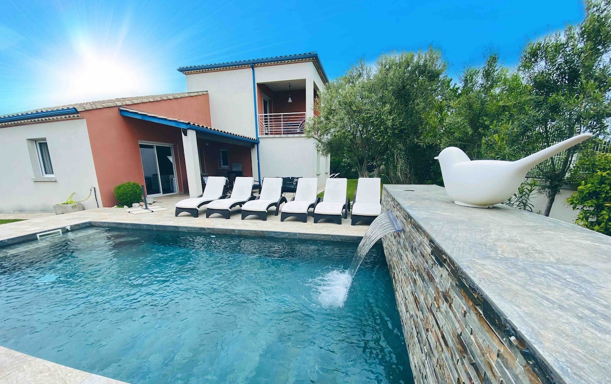 A spacious pool area features a modern swimming pool with a waterfall cascading into it. Six comfortable loungers are arranged around the pool, inviting relaxation. A well-maintained garden with olive trees is framed by the villa, which presents a clean and contemporary design.