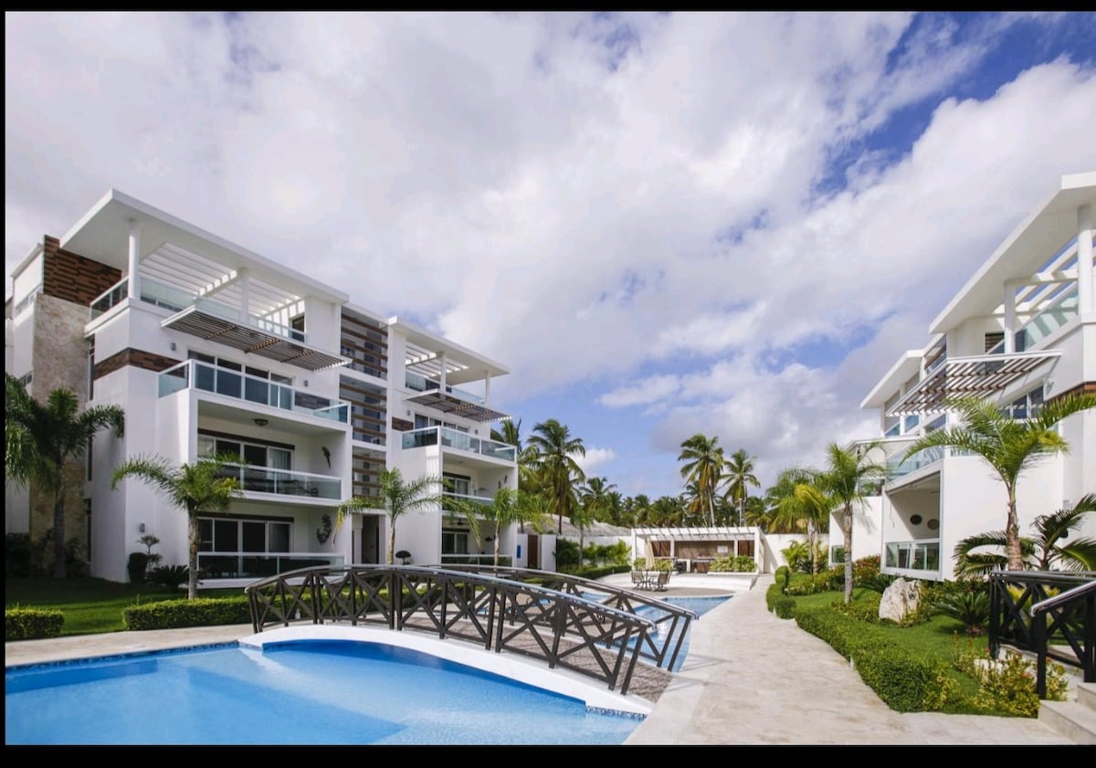 Two modern buildings with multiple balconies are visible against a serene sky. A bridge crosses over a blue swimming pool, bordered by tropical palm trees and manicured greenery, enhancing the outdoor space for relaxation.