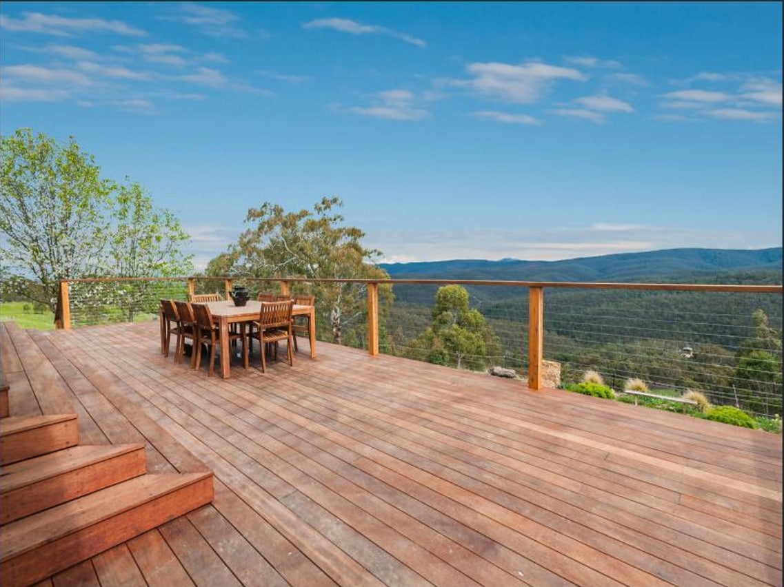 A spacious wooden deck is presented, featuring a dining table with six chairs positioned to enjoy expansive views of the surrounding landscape. Green trees and rolling hills are visible in the background, with blue skies enhancing the serene atmosphere.