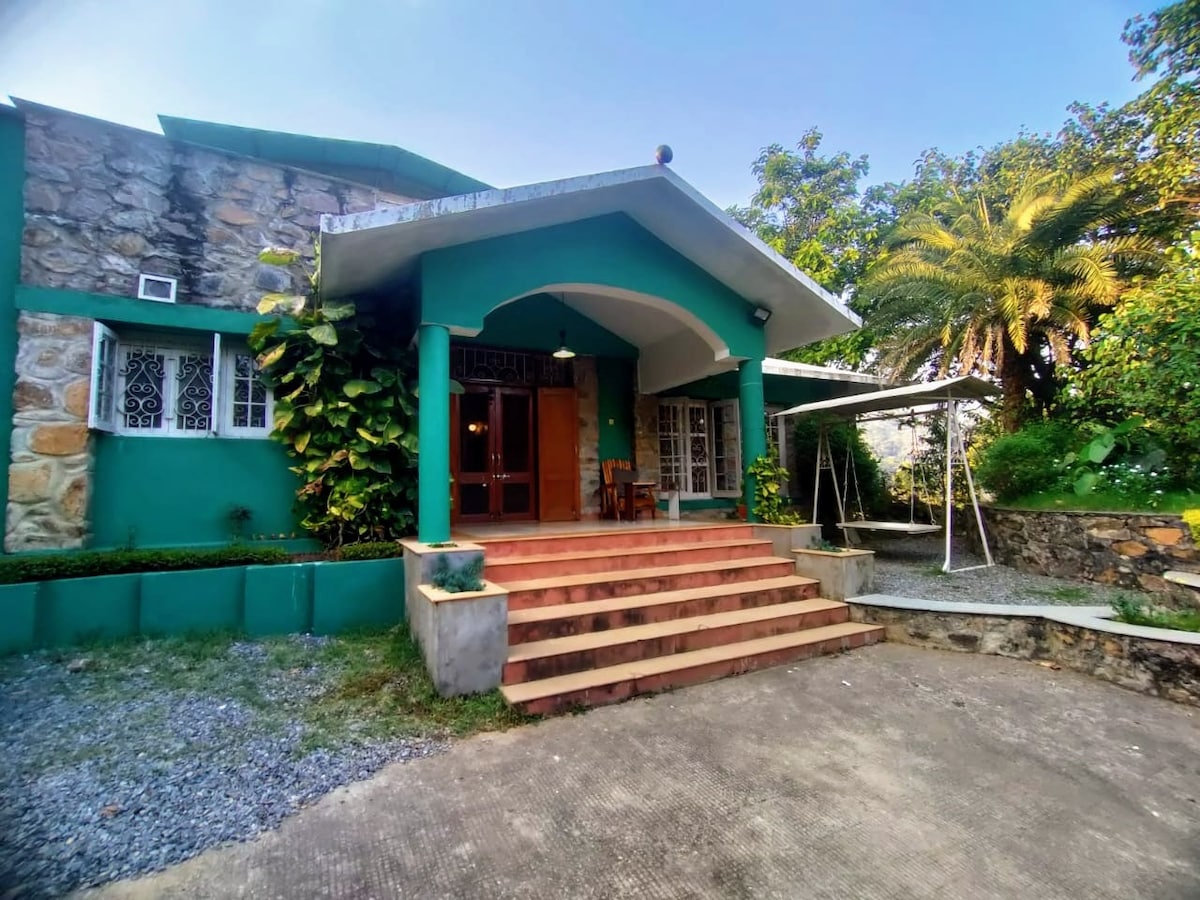 The entrance of the outhouse features a set of stone steps leading up to a green archway. Lush plants frame the entrance, while a palm tree is visible in the surrounding area. The front porch offers seating arrangements, inviting guests to relax outdoors.