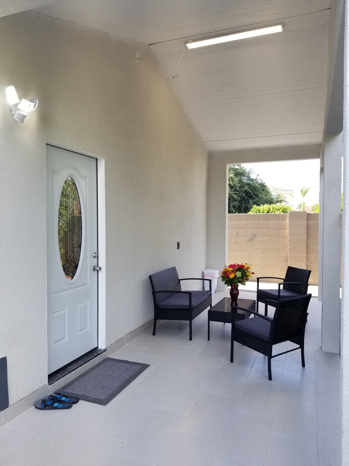 A covered entrance area is shown, featuring a white door with an oval window. Two black chairs and a small glass table are positioned on the tiled floor. A bouquet of flowers is placed on the table, and a mat is located near the door.