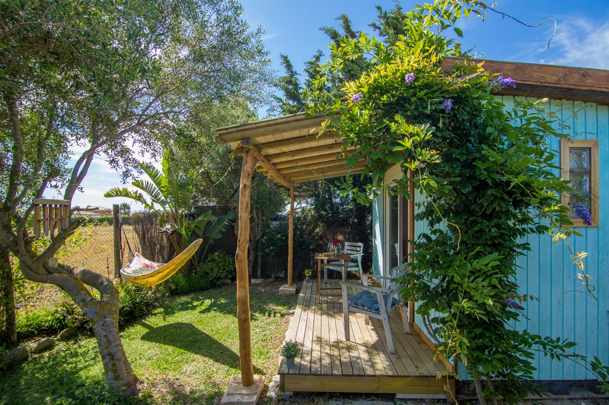 A wooden cabin features a covered porch surrounded by lush greenery. A yellow hammock hangs between the trees, and a small table with chairs is set on the wooden deck. Vibrant foliage adds to the inviting outdoor space.
