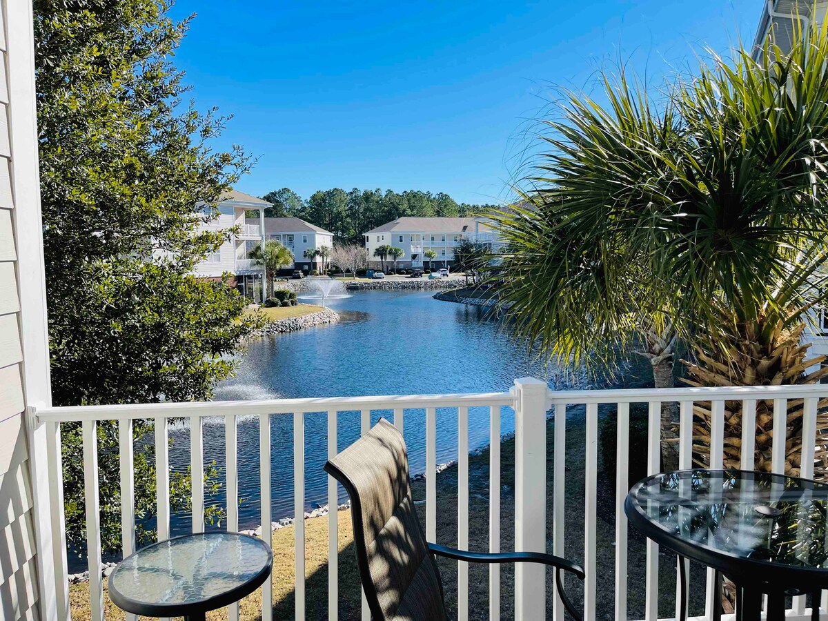 A serene outdoor view from a balcony overlooking a calm pond. Fountains can be seen gently bubbling in the water. Lush greenery frames the scene, with two chairs and a glass table positioned for outdoor relaxation.