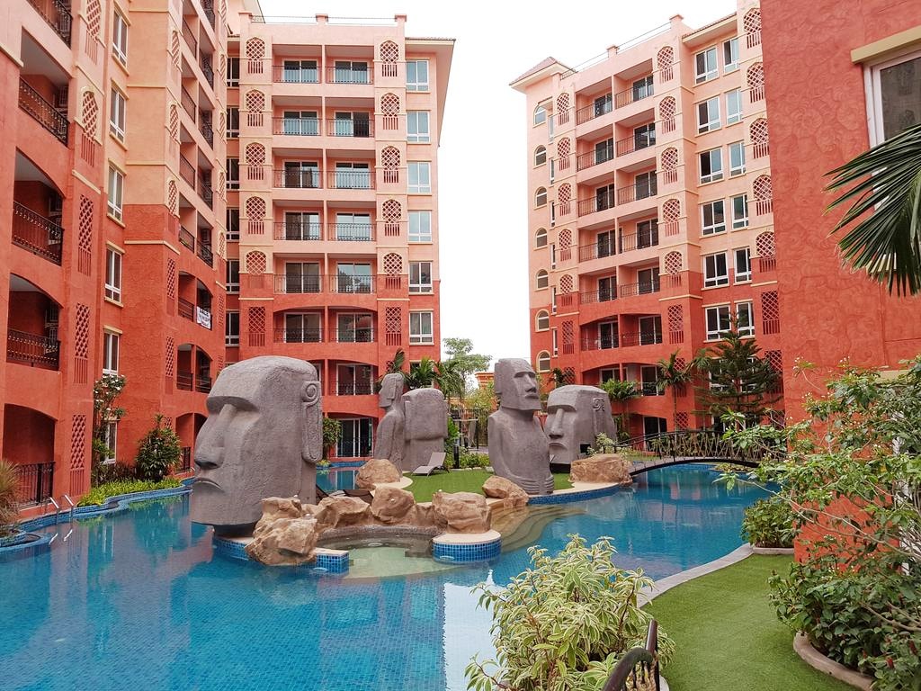 An inviting pool area features a unique arrangement of large stone sculptures surrounded by a well-maintained lawn. The vibrant red buildings and tiled walkways enhance the resort-like ambiance, while lush greenery provides an element of tranquility in the courtyard.