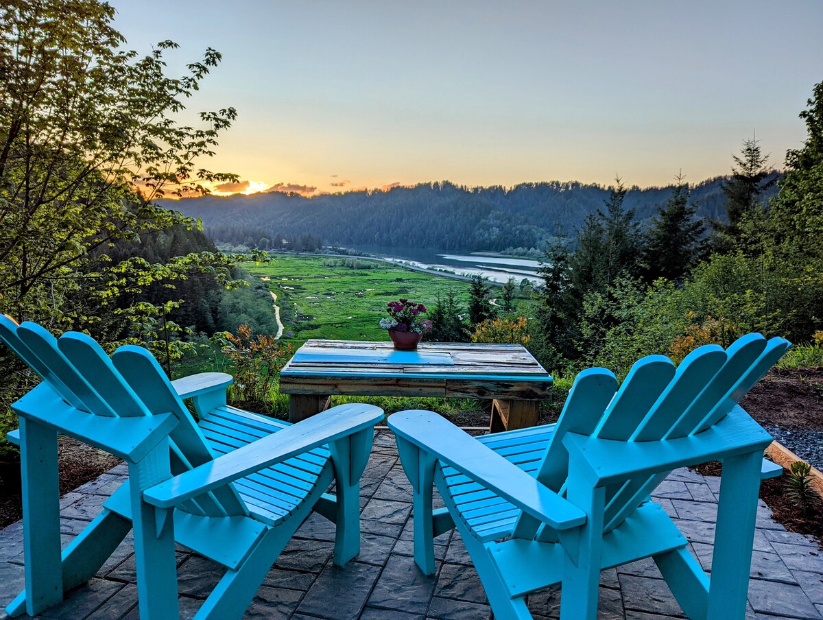 Two handcrafted Adirondack chairs with a turquoise finish are positioned around a rustic table, overlooking a lush valley. A small potted plant sits at the center of the table, with a stunning view of the sunset illuminating the landscape.
