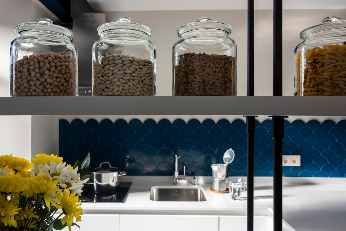 A modern kitchen is showcased, featuring a sleek countertop with a stainless-steel sink. Glass jars filled with various grains and pasta are arranged on a shelf above. A vibrant blue backsplash adds a pop of color, with a small pot of yellow flowers placed nearby.