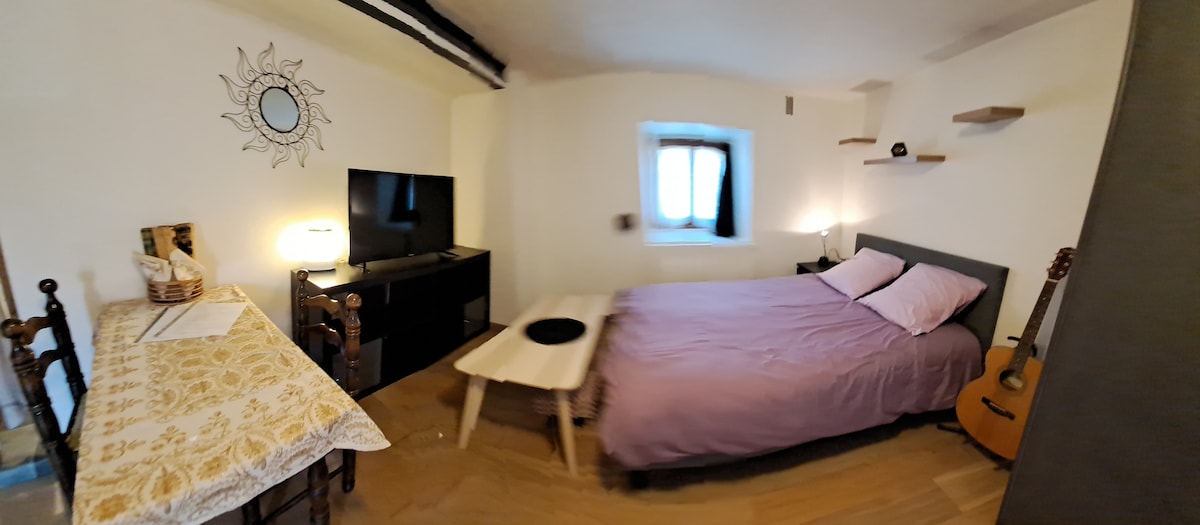 A cozy studio is displayed, featuring a spacious bed with a soft purple coverlet. A small dining table with seating for two is present, along with a television and shelves for storage. A guitar rests beside the bed, and a window allows natural light to enter.