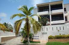 A modern building is shown with a well-maintained façade and large windows. A palm tree is positioned in the foreground, adding a tropical touch, while manicured greenery surrounds the entrance. Steps lead to the main entrance, emphasizing accessibility.