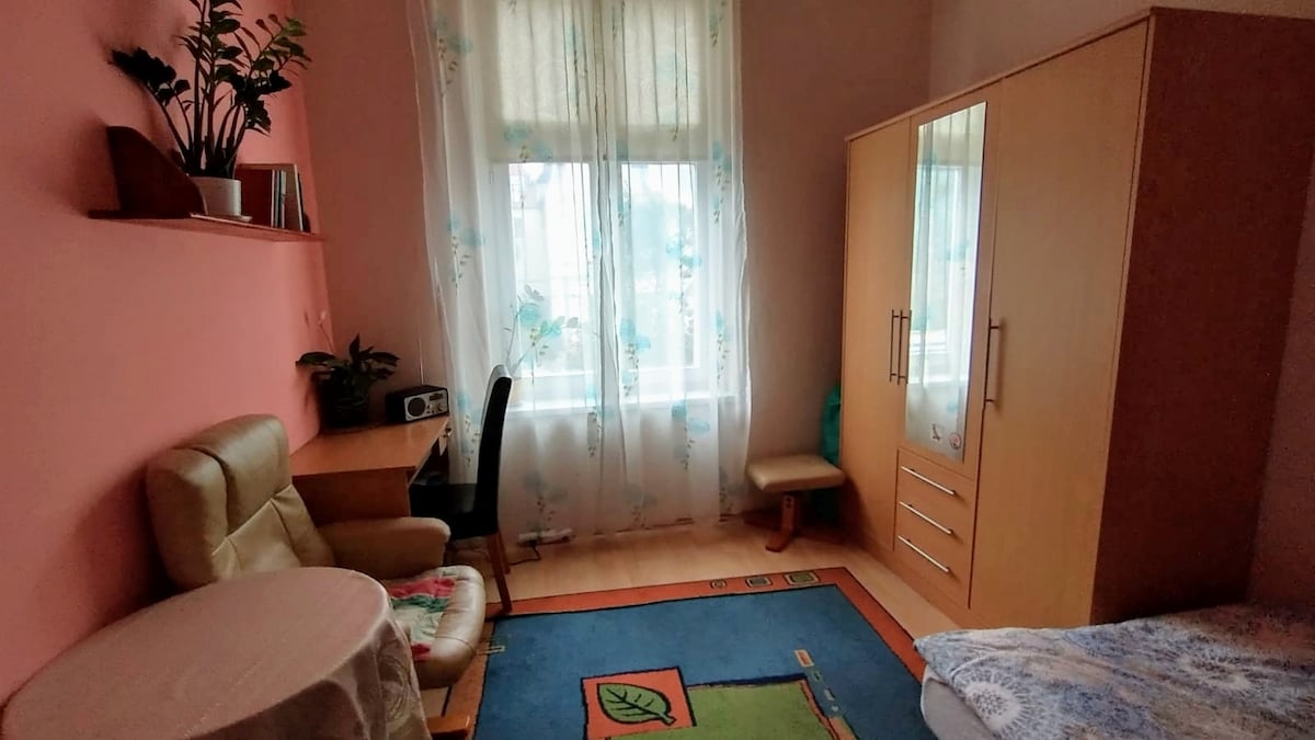 A bright room features a large window draped with sheer curtains, allowing natural light to fill the space. A small desk is paired with a chair, and a cozy armchair is positioned beside a round table. A wardrobe stands against one wall, with a colorful rug covering the floor.