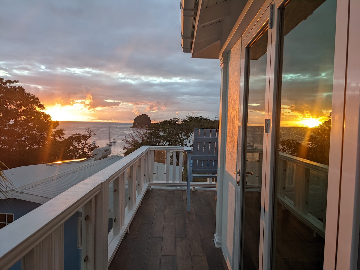 A serene private terrace is visible, showcasing panoramic views of the ocean at sunset. The scene features reflective glass doors, allowing natural light to illuminate the area. The horizon is painted with warm hues, while a distant rock formation enhances the coastal landscape.