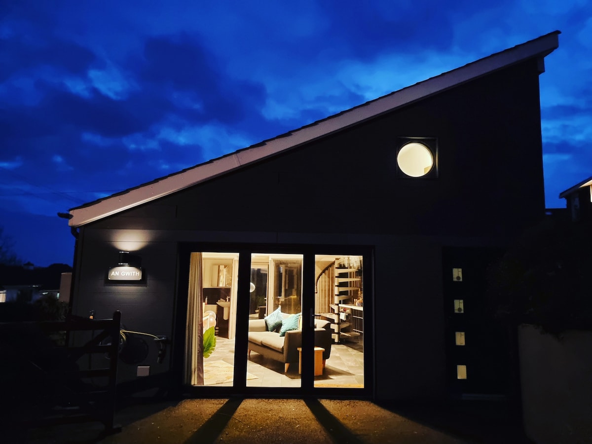 The exterior of An Gwith is presented at twilight, featuring a warm glow from the interior lights. A large window and bi-folding doors reveal glimpses of the lounge area, while a circular window adds architectural interest to the gable end.