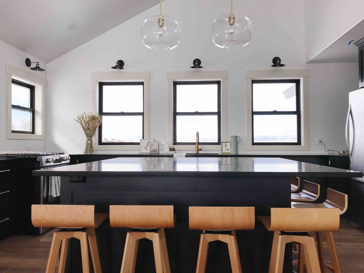 The kitchen features a large black granite island with seating for four. Natural light is provided by three windows, framed by white trim, with views of the surroundings. Modern pendant lights hang above the island, and sleek cabinetry adds to the contemporary look.