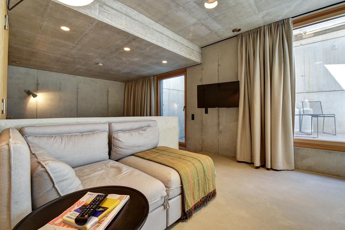 An open living space features a comfortable sofa bed draped with a striped throw. A small round table with a remote sits adjacent to it. Light filters through large windows with neutral curtains, complementing the exposed concrete walls and ceiling.
