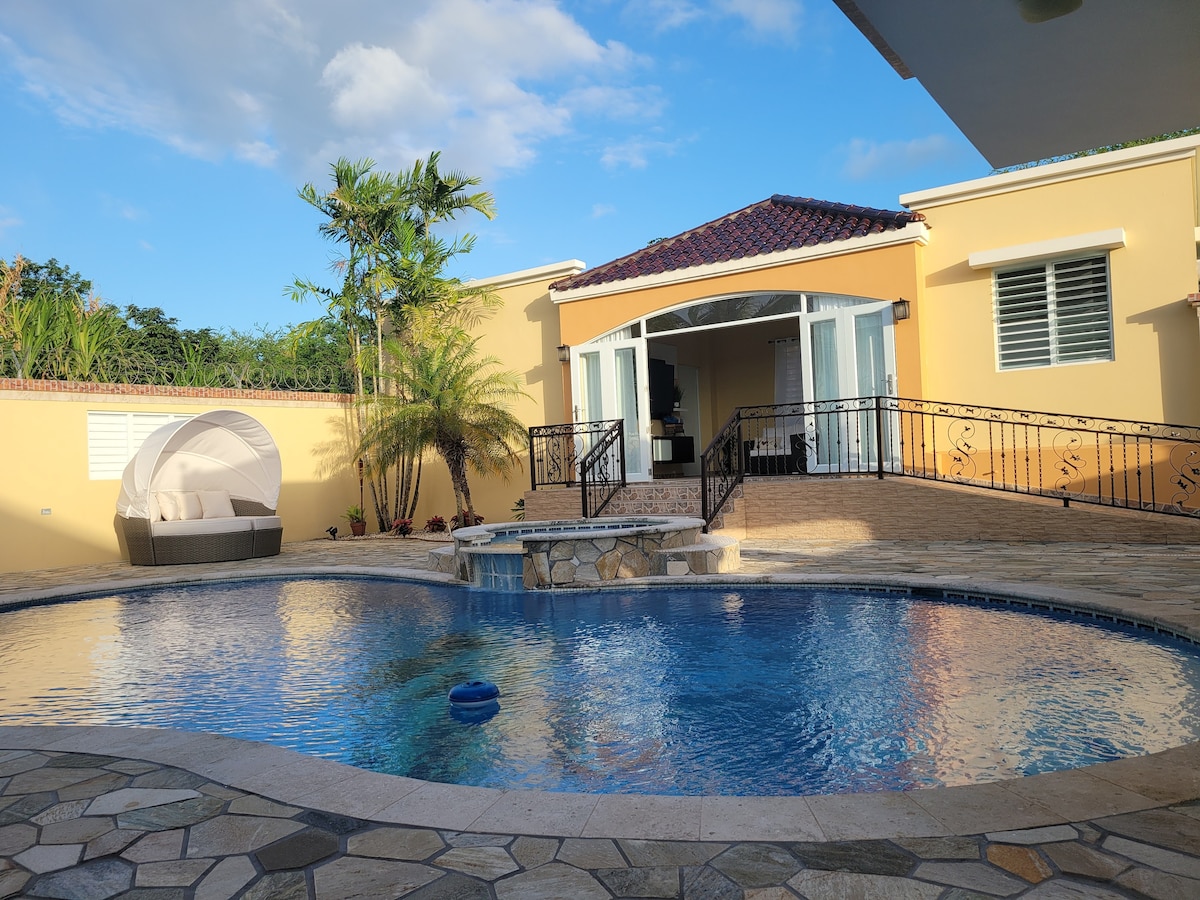 A serene outdoor area features a circular pool surrounded by natural stone. A charming gazebo sits nearby, providing shaded seating. Lush palm trees and vibrant landscaping enhance the setting, while a spacious villa is visible in the background, showcasing elegant architectural details.