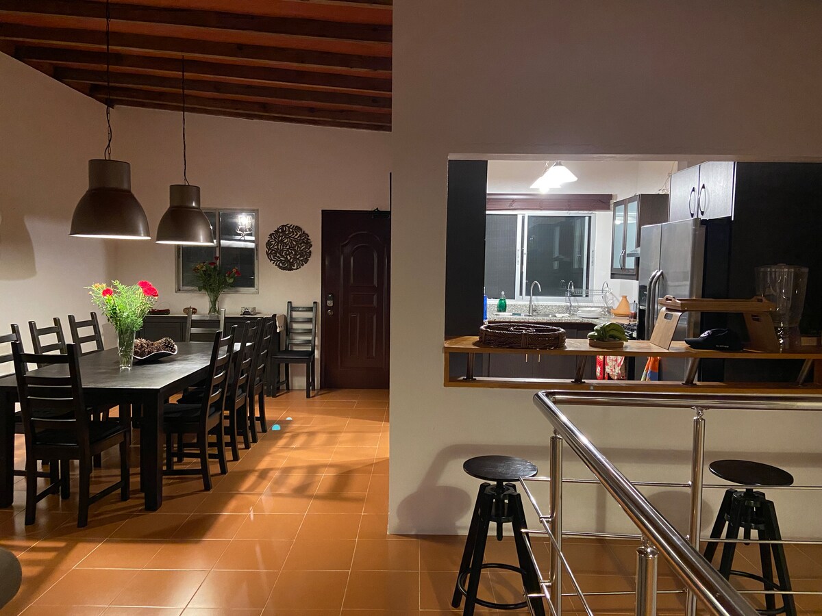 The spacious dining area features a large black table surrounded by six chairs, complemented by a flower vase. A modern kitchen is visible in the background, equipped with stainless steel appliances and a counter area with additional seating. Warm lighting enhances the inviting space.