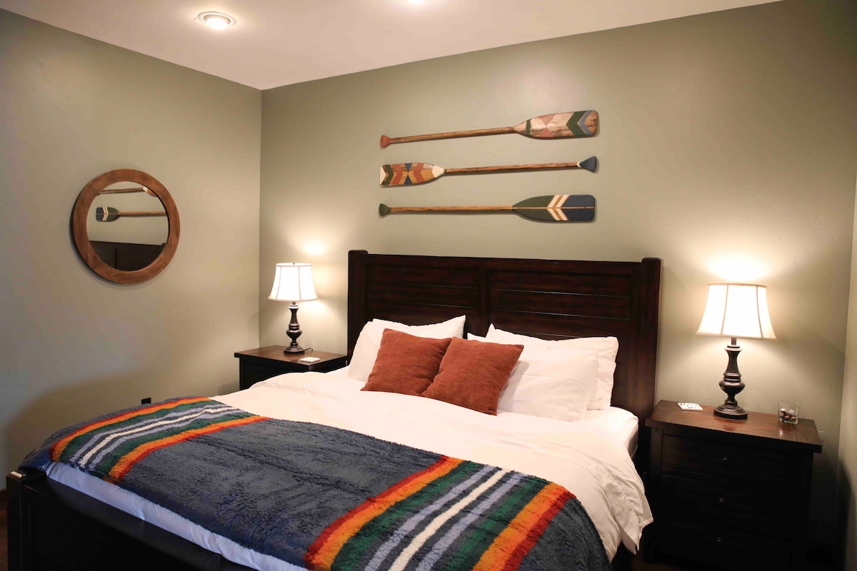 A cozy bedroom is furnished with a dark wood headboard and two bedside tables. Soft white linens are layered with a colorful striped blanket and decorative pillows. Two lamps provide gentle illumination. The walls feature a circular mirror and decorative oars arranged artfully.