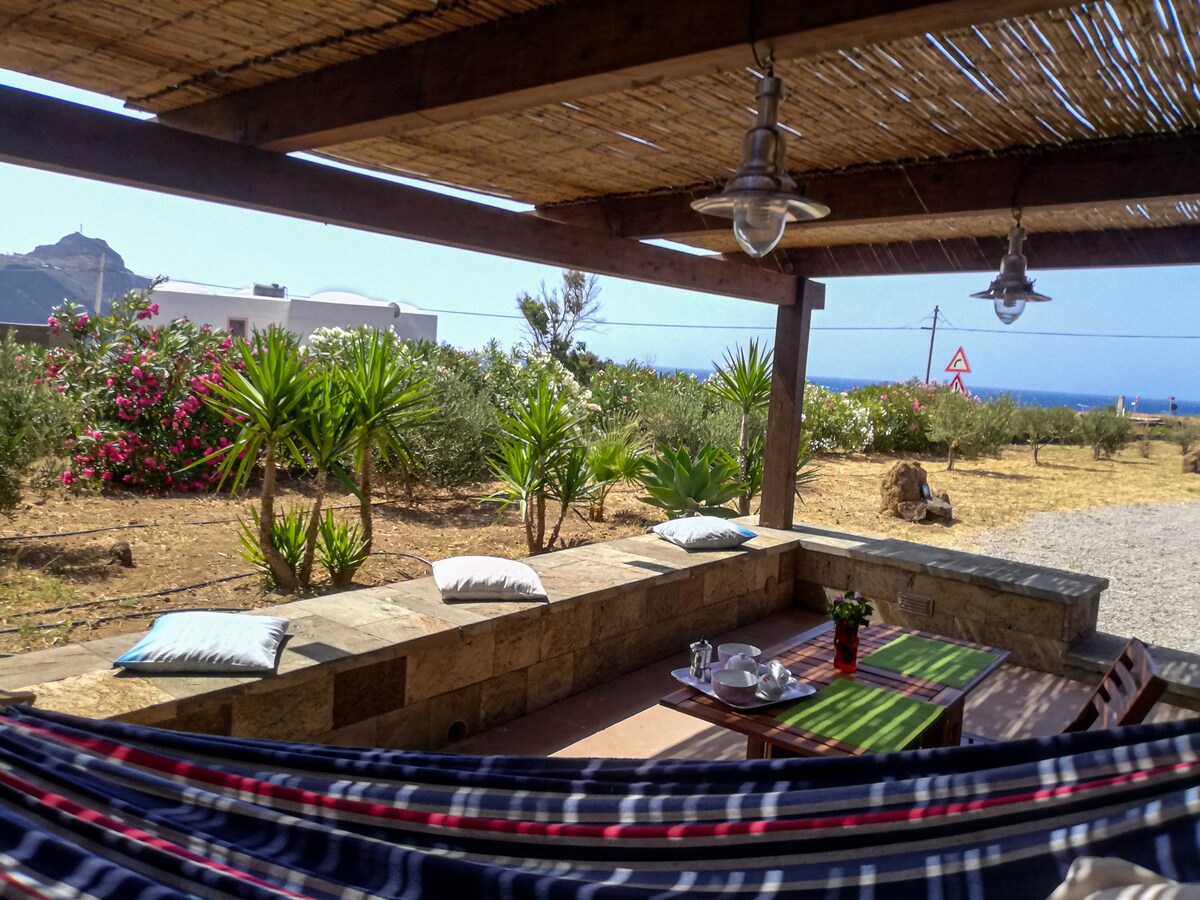 An outdoor veranda features a stone seating area surrounded by lush greenery and colorful flowers. A striped hammock is draped between wooden beams. A small table holds a vase with flowers, while the view extends toward the distant sea and a rocky landscape.