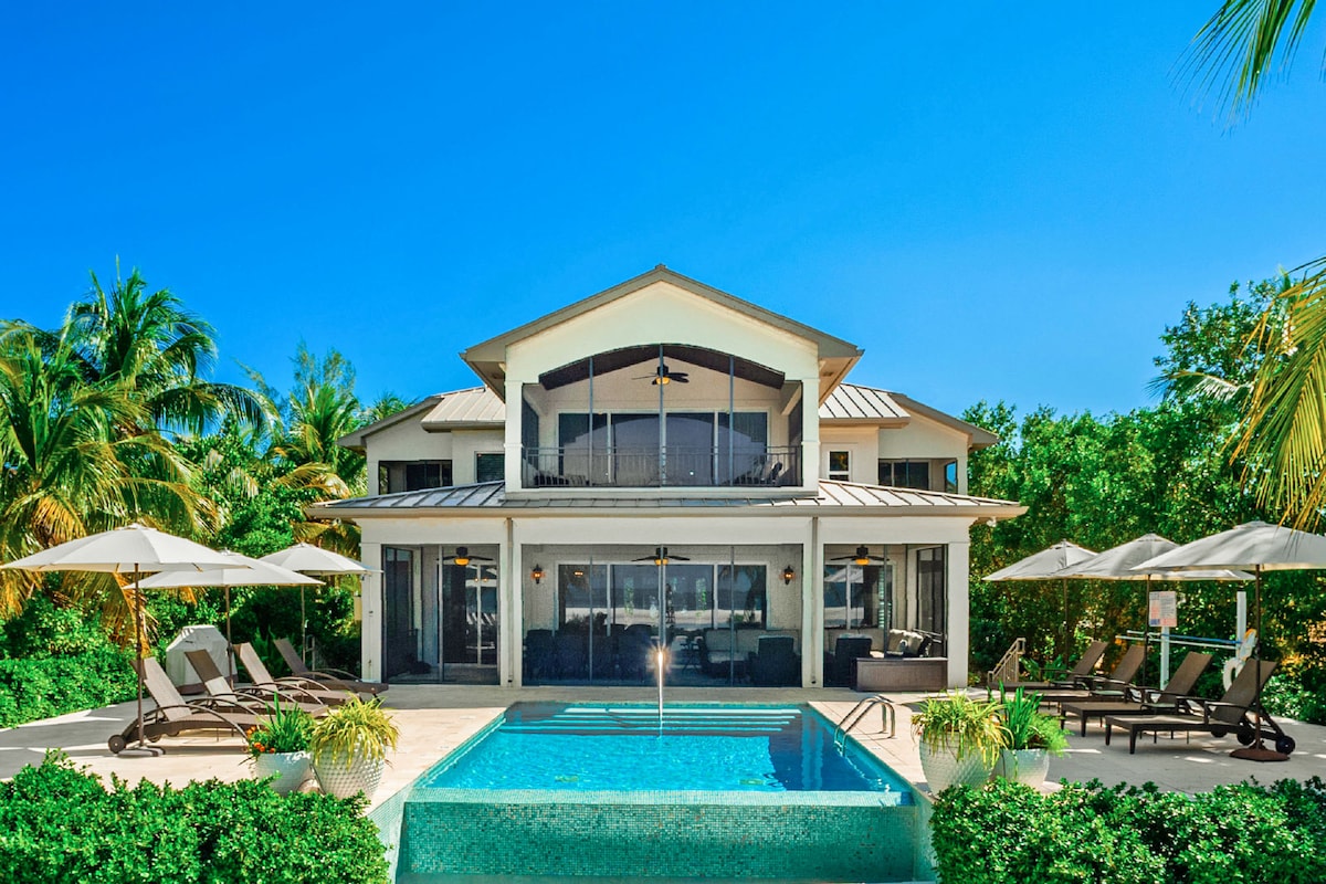 A spacious villa is showcased with a clear blue sky overhead and lush greenery surrounding the property. An inviting infinity pool is positioned in the foreground, flanked by sun loungers and shaded umbrellas, while large sliding glass doors provide a view of the interior.