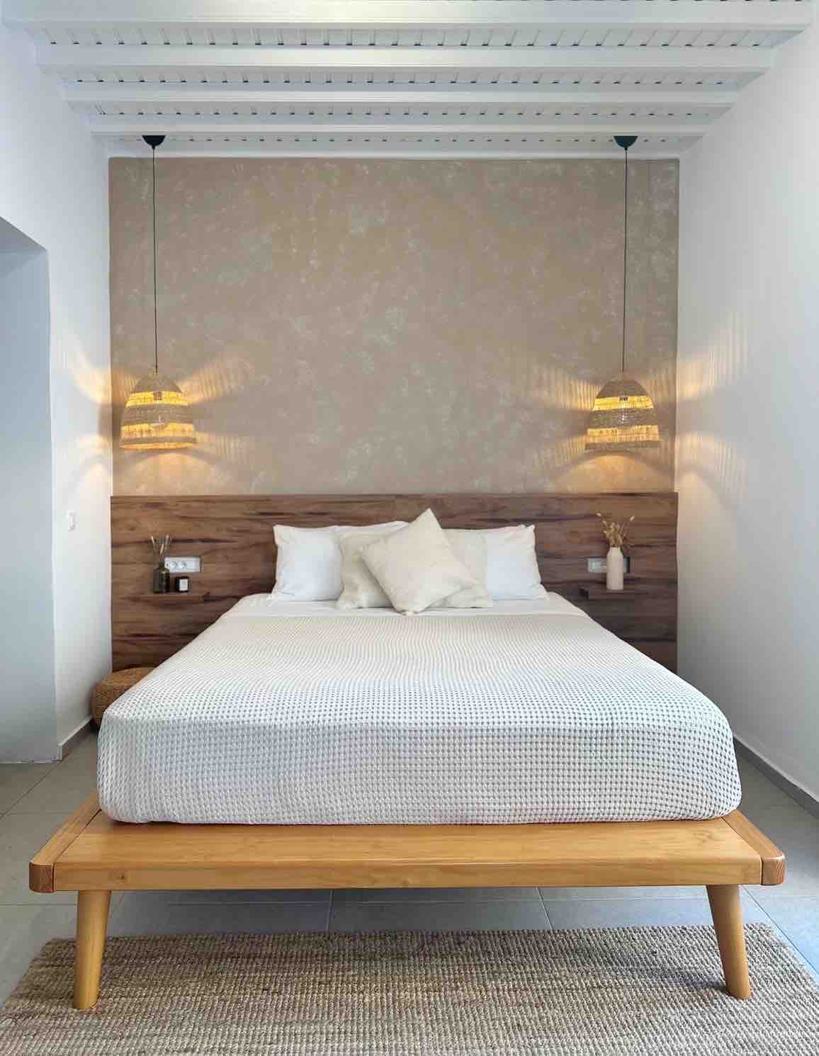 A cozy bedroom features a wooden bed frame topped with a light-colored woven blanket. Soft pillows are arranged against a textured wall, while two pendant lights hang above, providing gentle illumination. A natural fiber rug lies underneath, contributing to the room's warm atmosphere.