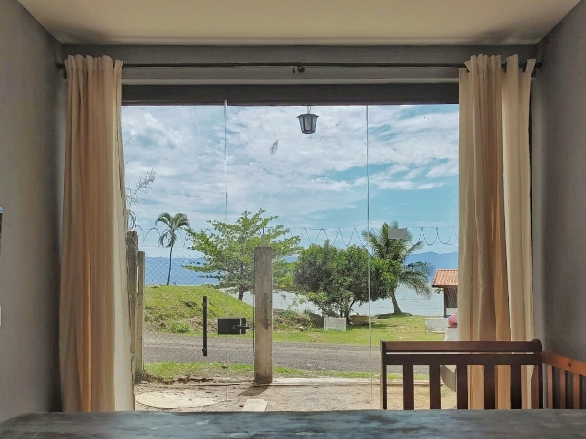 A wide opening presents a view of the ocean, framed by flowing curtains. Lush greenery and palm trees are visible in the foreground, alongside the tranquil water. The scene is brightened by natural light, contributing to a serene ambiance.