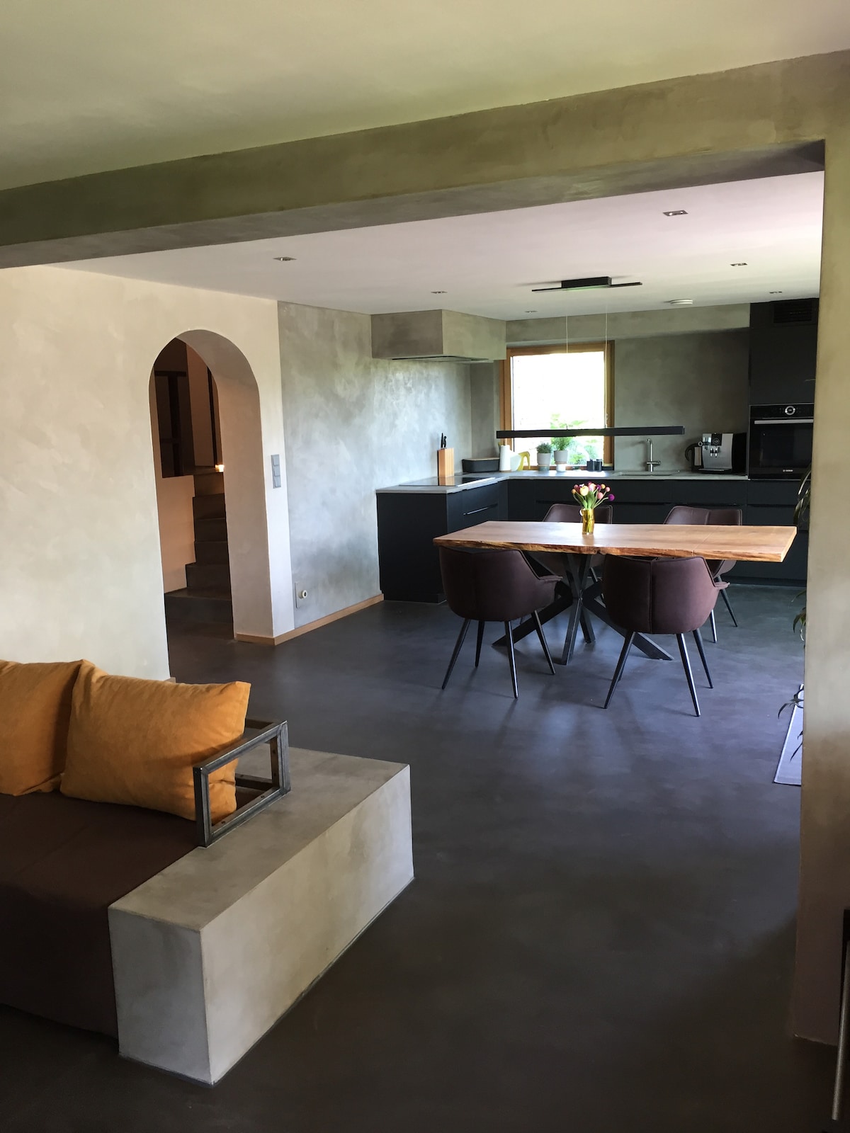 An open living space features a minimalist kitchen with modern black cabinetry and high-quality appliances. A dining table surrounded by four chairs is positioned next to the kitchen. A cozy seating area with an earthy color palette is visible, enhancing the overall spaciousness.